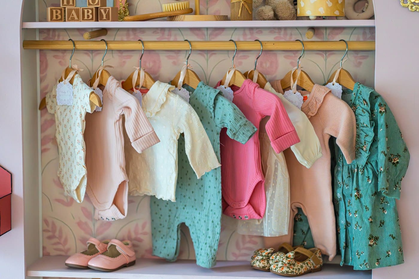 Pastel baby bodysuits and rompers on wooden hangers above small shoes